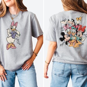 Pode incluir: Camiseta cinza mesclada com a Pata Daisy na frente e um grupo de personagens da Disney nas costas. Pata Daisy usa um top roxo e um la&ccedil;o rosa. A parte de tr&aacute;s apresenta Mickey Mouse, Minnie Mouse e Pluto.