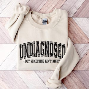 Może przedstawiać: Beżowa bluza z napisem "UNDIAGNOSED" w czarnym, uniwersyteckim kroju pisma. Poniżej znajduje się fraza "- BUT SOMETHING AIN'T RIGHT" w mniejszej, czarnej czcionce. Bluza jest złożona na jasnej powierzchni z fakturą drewna.