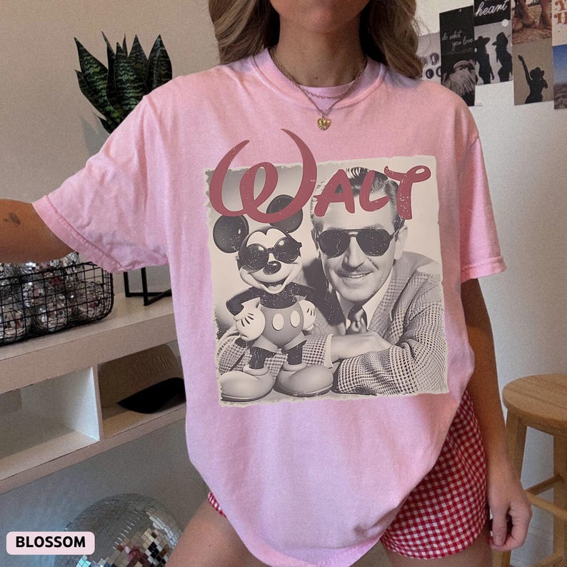 May include: A light pink short-sleeve t-shirt featuring a graphic print of a man in a suit and sunglasses standing next to a cartoon mouse wearing sunglasses. The text "WALT" is printed in a large, stylized red font above the graphic illustration.