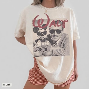May include: An ivory short-sleeve t-shirt featuring a graphic print of Mickey Mouse and Walt Disney, both wearing sunglasses. The word "Walt" is printed in large, distressed red script above the figures. The shirt has a crew neckline.