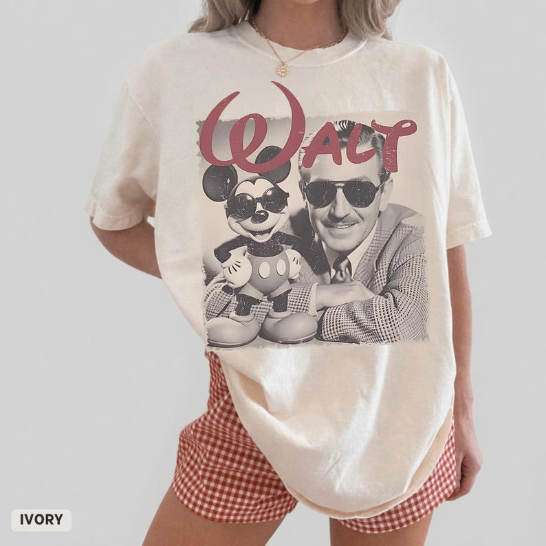 May include: An ivory short-sleeve t-shirt featuring a graphic print of Mickey Mouse and Walt Disney, both wearing sunglasses. The word "Walt" is printed in large, distressed red script above the figures. The shirt has a crew neckline.