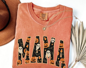May include: Orange Comfort Colors sweatshirt with the word "MAMA" in a black and orange Halloween-themed design. The design includes jack-o'-lanterns, ghosts, and candy corn. A gold necklace is draped across the shirt.