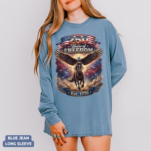 May include: A blue jean long-sleeve shirt featuring a graphic with a horse, eagle wings, and the text "250 Years of Freedom Est. 1776". The design incorporates red, white, and blue elements.
