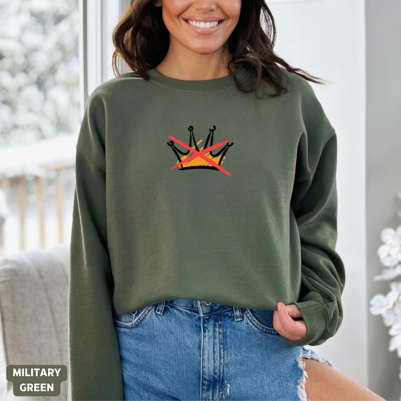 Puede incluir: Sudadera verde militar con un dise&ntilde;o de corona tachada. La sudadera de cuello redondo es de color liso con un peque&ntilde;o gr&aacute;fico en la parte superior izquierda del pecho. El texto "MILITARY GREEN" est&aacute; en la parte inferior izquierda de la imagen.