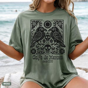 Puede incluir: Camiseta verde musgo con un dise&ntilde;o gr&aacute;fico negro que muestra dos cuervos, calaveras, flores y el texto "Golfo de Mexico Desde 1550". El dise&ntilde;o est&aacute; centrado en la parte delantera.