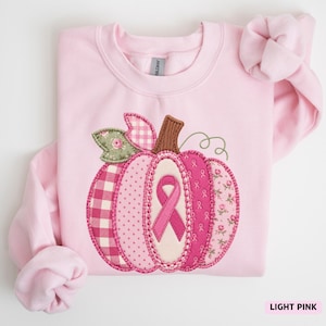 May include: A light pink crewneck sweatshirt featuring a large pumpkin applique on the front. The pumpkin is constructed from various pink fabric patches, including gingham, floral, and breast cancer awareness ribbon patterns, with a brown stem.