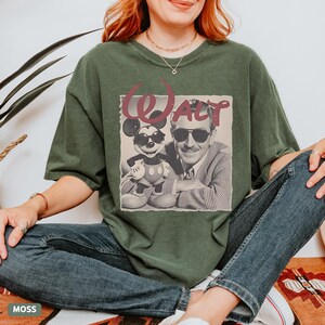 May include: A dark olive green short-sleeve t-shirt featuring a graphic print of Walt Disney and Mickey Mouse both wearing sunglasses. The text "Walt" is printed in a large, stylized maroon font above the black and white image of the pair.