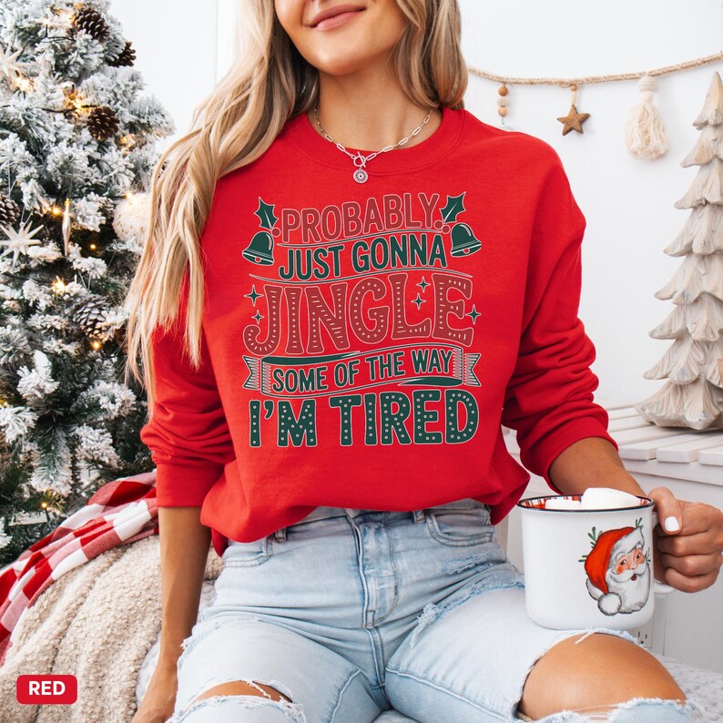 May include: A red Christmas sweatshirt with the text "Probably Just Gonna Jingle Some of the Way I'm Tired" in white and green. The sweatshirt is long-sleeved and features a crew neck. A white mug with a Santa Claus graphic is held in the hand.