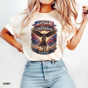 May include: Ivory-colored t-shirt featuring a patriotic design. The graphic includes an eagle with outstretched wings, a horse, and the text "250 Years of Freedom" above a banner that reads "Est. 1776". The design incorporates red, white, and blue elements.