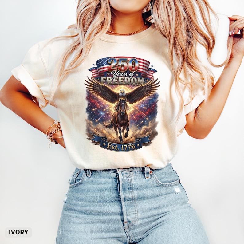 May include: Ivory-colored t-shirt featuring a patriotic design. The graphic includes an eagle with outstretched wings, a horse, and the text "250 Years of Freedom" above a banner that reads "Est. 1776". The design incorporates red, white, and blue elements.