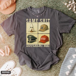 Puede incluir: Camiseta gris oscuro con el texto "SAME SHIT DIFFERENT HAT" y cuatro imágenes de sombreros. Las imágenes incluyen una capucha blanca, una gorra de béisbol beige, un sombrero militar negro y una gorra de béisbol roja.