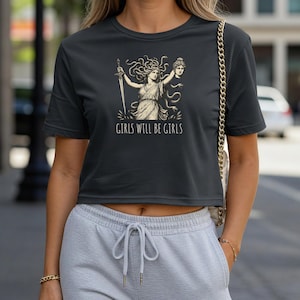 May include: Dark gray crop top featuring a graphic of a woman with a sword and a severed head, with the text "GIRLS WILL BE GIRLS" in white. The design is in a vintage style.