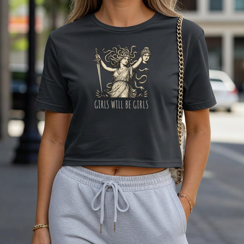 May include: Dark gray crop top featuring a graphic of a woman with a sword and a severed head, with the text "GIRLS WILL BE GIRLS" in white. The design is in a vintage style.