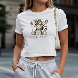 May include: White crop top featuring a graphic of a woman with a sword and a severed head, with the text "GIRLS WILL BE GIRLS" in black. The shirt has short sleeves and a crew neck.