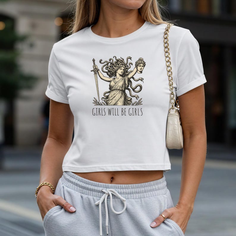 May include: White crop top featuring a graphic of a woman with a sword and a severed head, with the text "GIRLS WILL BE GIRLS" in black. The shirt has short sleeves and a crew neck.
