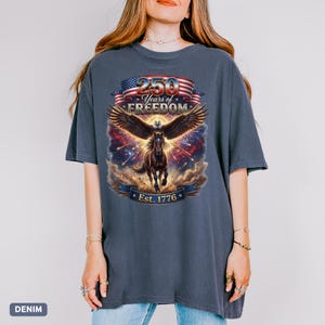 May include: A denim-colored t-shirt features a graphic celebrating 250 Years of Freedom. The design includes an eagle with outstretched wings, a running horse, and the text "Est. 1776" on a banner, with an American flag.