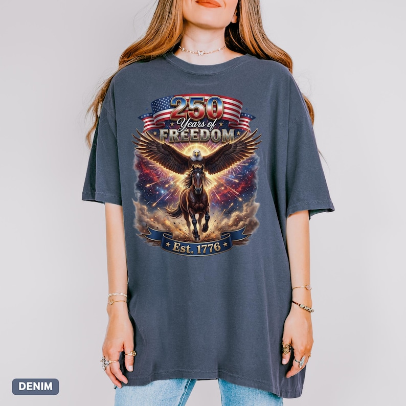 May include: A denim-colored t-shirt features a graphic celebrating 250 Years of Freedom. The design includes an eagle with outstretched wings, a running horse, and the text "Est. 1776" on a banner, with an American flag.