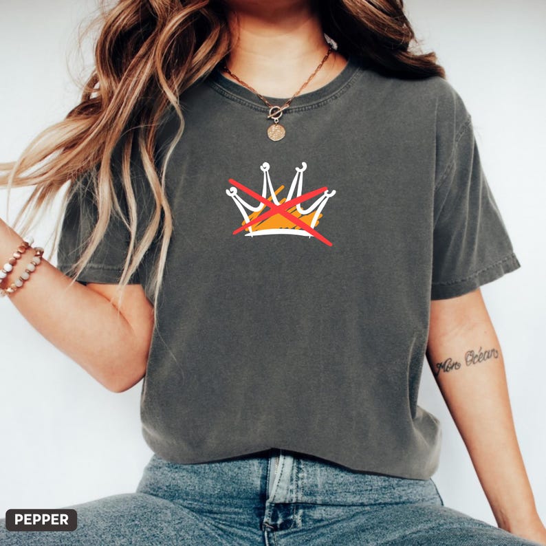 May include: Dark grey t-shirt featuring a graphic of a crown with a red line through it. The crown is white and yellow. The shirt is a crew neck and has short sleeves.