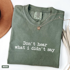May include: A moss green t-shirt with the phrase "Don't hear what I didn't say" printed in white. A gold necklace with a pendant rests on the shirt. The shirt is folded and has a Comfort Colors tag.