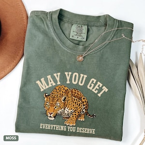 May include: A moss green t-shirt with the text "MAY YOU GET EVERYTHING YOU DESERVE" and a detailed illustration of a leopard. A gold necklace and a brown hat are also visible.