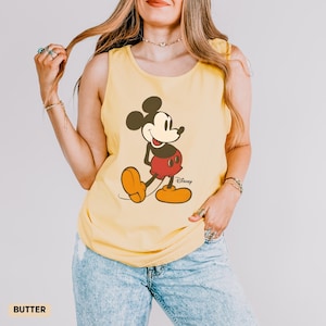 Puede incluir: Una camiseta sin mangas de color amarillo mantequilla con un estampado gr&aacute;fico de Mickey Mouse con sus cl&aacute;sicos pantalones cortos rojos y zapatos amarillos. El personaje est&aacute; centrado en la parte delantera con la palabra Disney en negro.