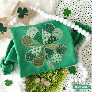 May include: A green St. Patrick's Day hat with a large, patchwork shamrock design. The shamrock is made of various green fabrics. The hat is surrounded by decorative elements, including shamrock-shaped cutouts and a white pom-pom garland.