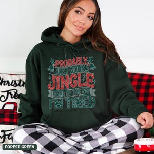May include: Forest green hoodie with the text "PROBABLY JUST GONNA JINGLE SOME OF THE WAY I'M TIRED" in red and white. The hoodie has a drawstring hood and a front pocket. The model is wearing black and white plaid pajama pants.