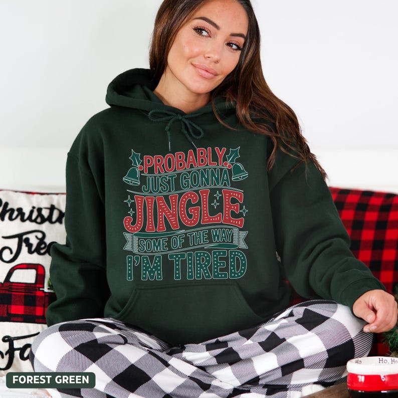 May include: Forest green hoodie with the text "PROBABLY JUST GONNA JINGLE SOME OF THE WAY I'M TIRED" in red and white. The hoodie has a drawstring hood and a front pocket. The model is wearing black and white plaid pajama pants.