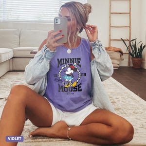 K&ouml;nnte beinhalten: Ein violettes Tanktop mit einem Minnie Mouse-Motiv. Die Grafik enth&auml;lt den Text "MINNIE MOUSE" in einem kreisf&ouml;rmigen Design mit Minnie Mouse in einem gepunkteten Kleid. Das Tanktop wird mit wei&szlig;en Shorts und einer Jeansjacke getragen.