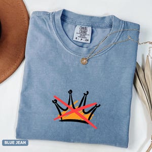 May include: Blue jean colored t-shirt with a graphic of a crown crossed out in red. The shirt has a crew neck and short sleeves. A gold necklace with a pendant is draped across the shirt.