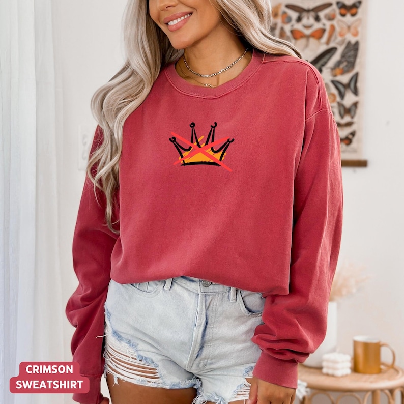 May include: Crimson sweatshirt with a crew neck and long sleeves. The front features a graphic of a crown with a red line through it, in black, yellow, and red. The text "CRIMSON SWEATSHIRT" is at the bottom.