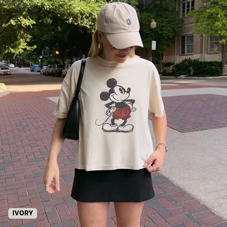 K&ouml;nnte beinhalten: Cremefarbenes T-Shirt mit einem Mickey Mouse-Motiv in Schwarz, Rot und Wei&szlig;. Das Shirt hat kurze &Auml;rmel und eine lockere Passform. Das Wort "IVORY" ist unten links im Bild aufgedruckt. Die Person tr&auml;gt eine beige Kappe und einen schwarzen Rock.
