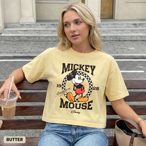 May include: Pale yellow short-sleeved t-shirt with a Mickey Mouse graphic. The design includes the words "Mickey Mouse" and the year "1928" within a black and white chequered circle. The shirt is worn with blue jeans.