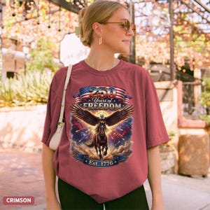 May include: Crimson t-shirt with a graphic featuring a horse with eagle wings, the American flag, and the text "250 Years of Freedom Est. 1776". The shirt is a relaxed fit.