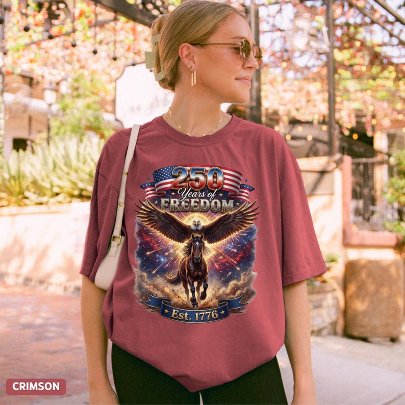 May include: Crimson t-shirt with a graphic featuring a horse with eagle wings, the American flag, and the text "250 Years of Freedom Est. 1776". The shirt is a relaxed fit.