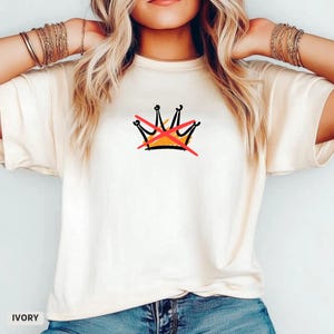 May include: Ivory-colored t-shirt featuring a hand-drawn crown design with a red 'X' through it. The graphic is in black, orange, and red. The shirt has short sleeves and a crew neck. The word "IVORY" is printed at the bottom left.