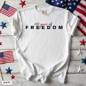 Peut inclure: T-shirt blanc avec l'inscription "250 years of FREEDOM" en noir, rouge et bleu. Le t-shirt est entouré de drapeaux américains et de décorations en forme d'étoiles rouges, blanches et bleues.