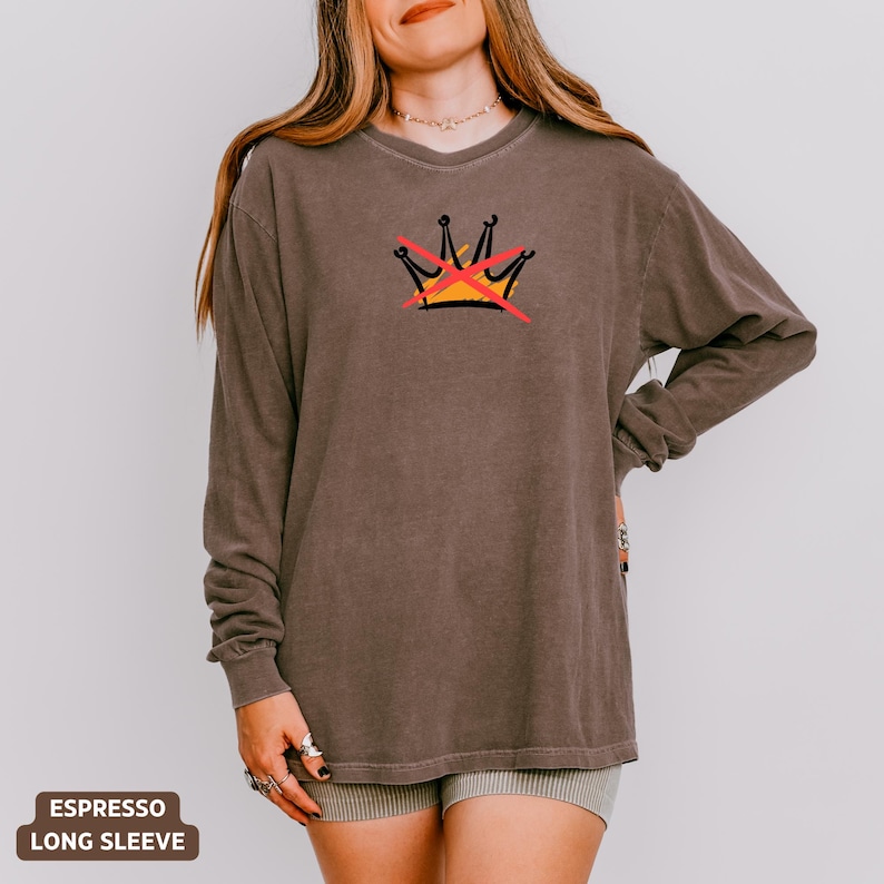 May include: Espresso-colored long-sleeve shirt with a graphic of a crown crossed out in red. The crown is yellow and black. The shirt is a solid color and has a relaxed fit.
