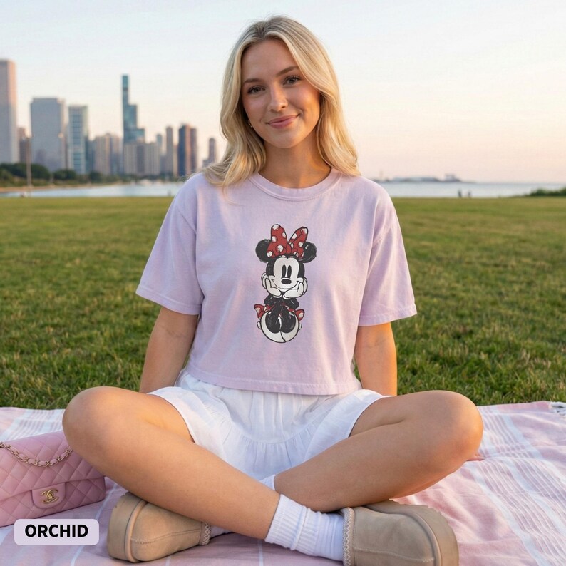 K&ouml;nnte beinhalten: Ein helllila Crop-Top mit einem Minnie Mouse-Motiv, das eine rote Schleife und schwarz-wei&szlig;e Details aufweist. Dazu wird ein wei&szlig;er Rock getragen. Das Wort "ORCHID" ist unten links aufgedruckt.