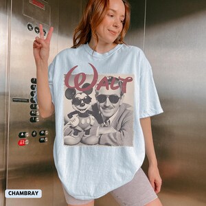 May include: A light blue t-shirt featuring a graphic print of Walt Disney and Mickey Mouse, both wearing sunglasses. The word "Walt" is printed in a large, stylized maroon font above the black and white image of the two characters.