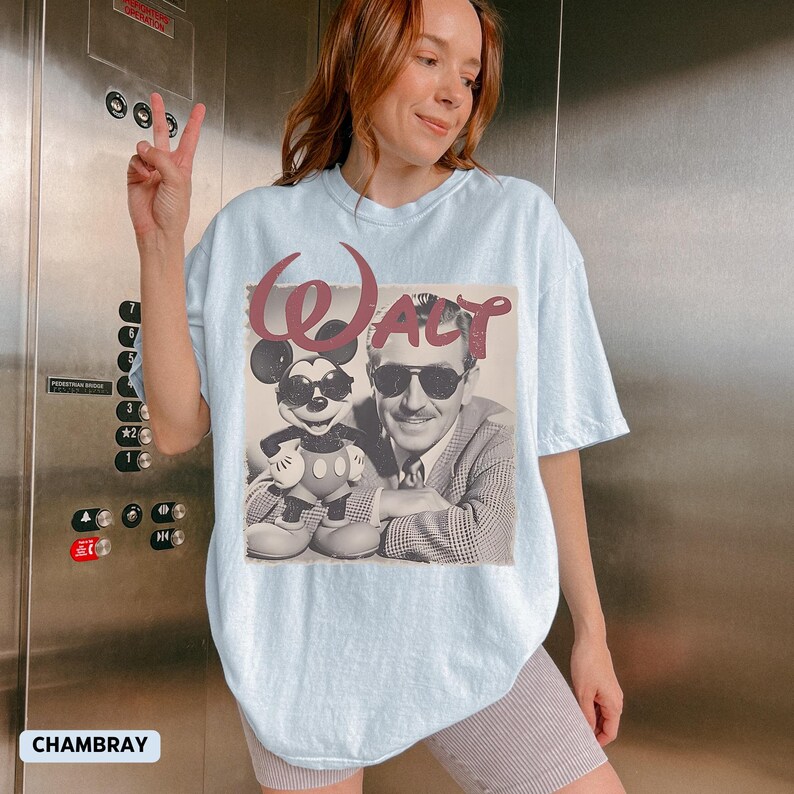 May include: A light blue t-shirt featuring a graphic print of Walt Disney and Mickey Mouse, both wearing sunglasses. The word "Walt" is printed in a large, stylized maroon font above the black and white image of the two characters.