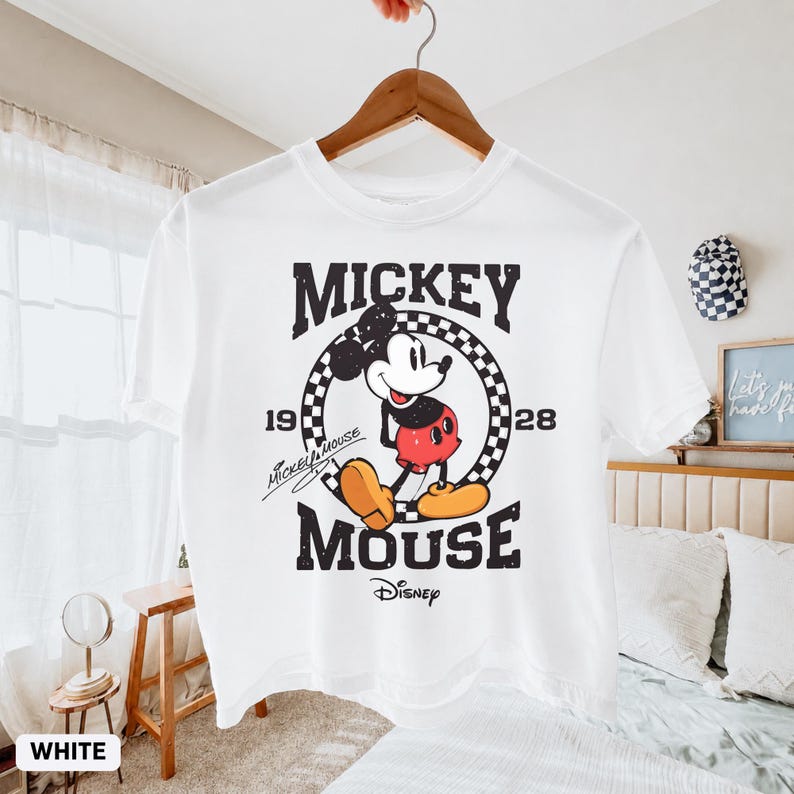 May include: White t-shirt with a vintage Mickey Mouse graphic. The design includes the words "Mickey Mouse" and "Disney" along with the year "1928". The shirt is suspended from a wooden hanger.