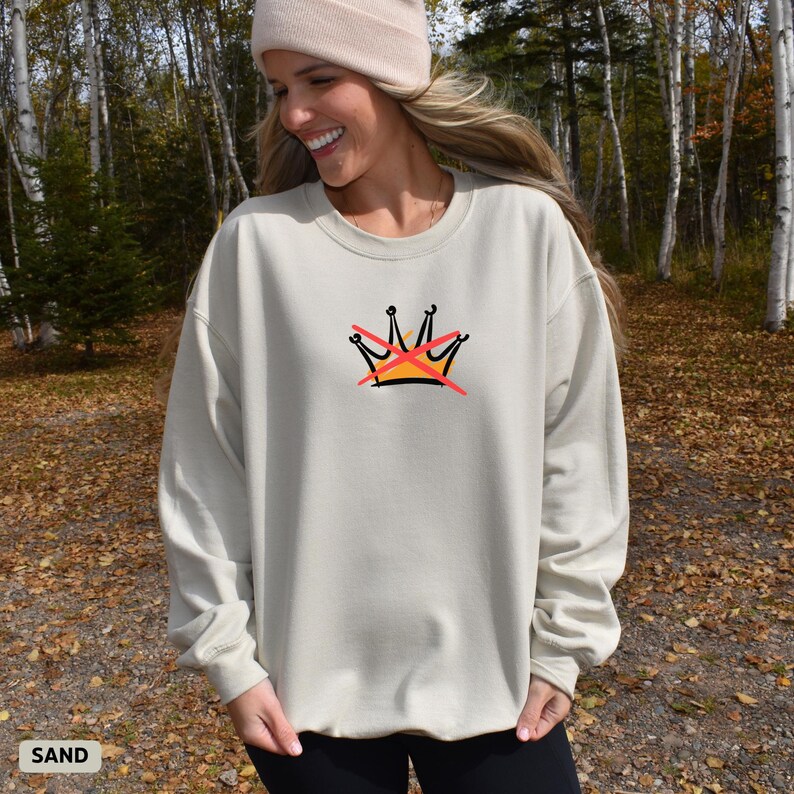 Puede incluir: Sudadera color arena con un gr&aacute;fico de corona tachada con l&iacute;neas rojas. La corona est&aacute; delineada en negro, con detalles en amarillo y naranja. La sudadera es de color neutro, adecuada para varios conjuntos.