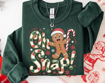 Könnte beinhalten: Forstgrünes Sweatshirt mit dem Text "Oh Snap!" und einem Lebkuchenmann mit Weihnachtsmütze und Zuckerstange. Das Design hat rote, weiße und grüne Akzente, ideal für die Weihnachtszeit.