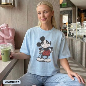 K&ouml;nnte beinhalten: Hellblaues T-Shirt mit einem Mickey Mouse-Cartoon-Aufdruck in Schwarz, Wei&szlig; und Rot. Die Comicfigur ist in einer klassischen Pose dargestellt. Das T-Shirt wird mit einer blauen Jeans kombiniert. Das Wort "CHAMBRAY" ist sichtbar.