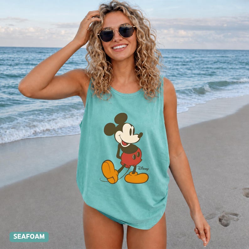Puede incluir: Una camiseta sin mangas de color verde espuma de mar con un estampado gr&aacute;fico de Mickey Mouse con sus caracter&iacute;sticos pantalones cortos rojos y zapatos amarillos. El personaje aparece en una pose cl&aacute;sica con el logo de Disney en negro.