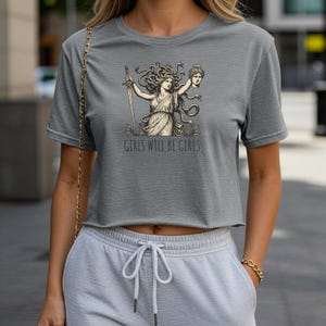 May include: Gray crop top featuring a Medusa graphic holding a sword and a severed head, with the text "GIRLS WILL BE GIRLS" below the graphic. The shirt is made of a soft material.