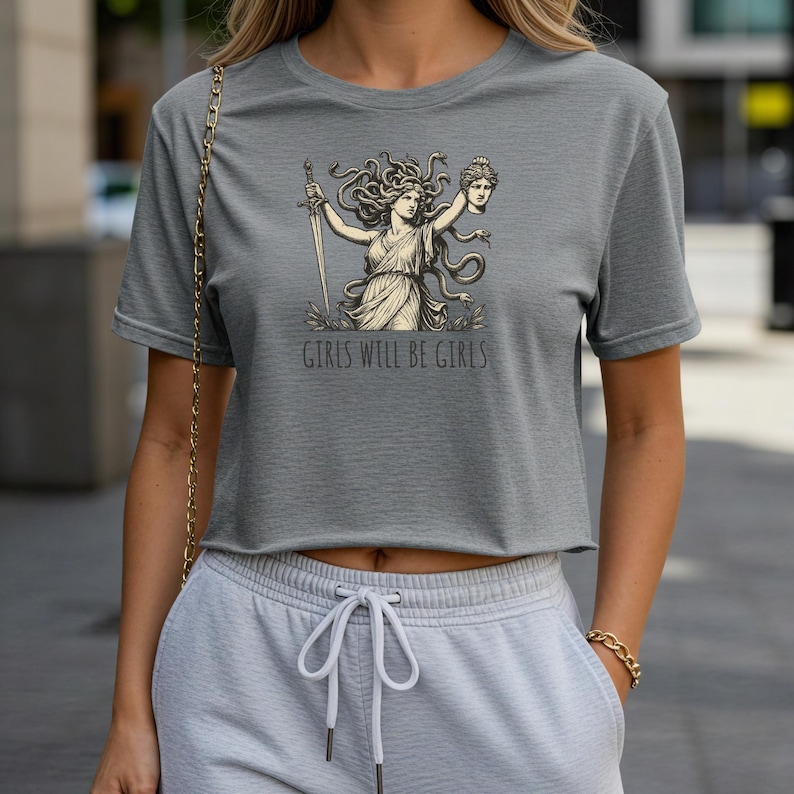 May include: Gray crop top featuring a Medusa graphic holding a sword and a severed head, with the text "GIRLS WILL BE GIRLS" below the graphic. The shirt is made of a soft material.