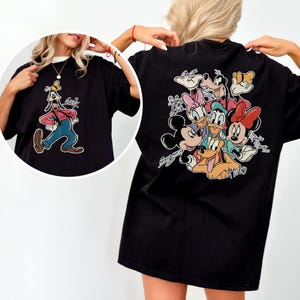 Pode incluir: Camiseta preta com uma imagem de desenho animado do Pateta na frente e um grupo de personagens da Disney nas costas. O gr&aacute;fico do Pateta mostra-o com uma camisa vermelha, cal&ccedil;as azuis e um chap&eacute;u amarelo. As costas apresentam Mickey, Minnie, Donald, Margarida, Pluto e Pateta.