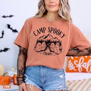 May include: A peach-coloured t-shirt with the words "CAMP SPOOKY" in black, above a graphic of ghosts around a campfire, mountains, and a cabin. The shirt is paired with denim shorts.